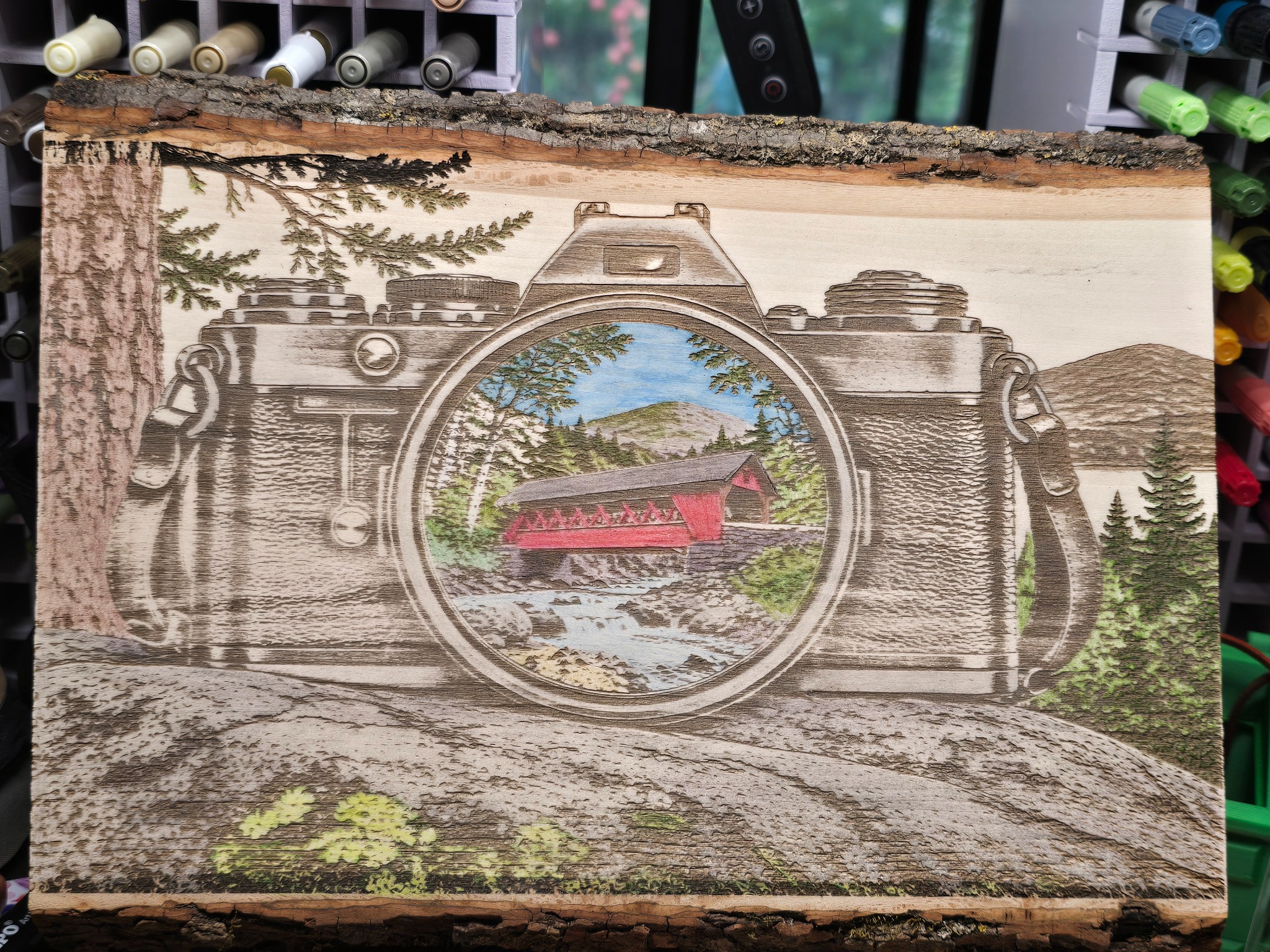 Wooden camera-shaped engraved artwork with a colorful covered bridge scene inside the lens